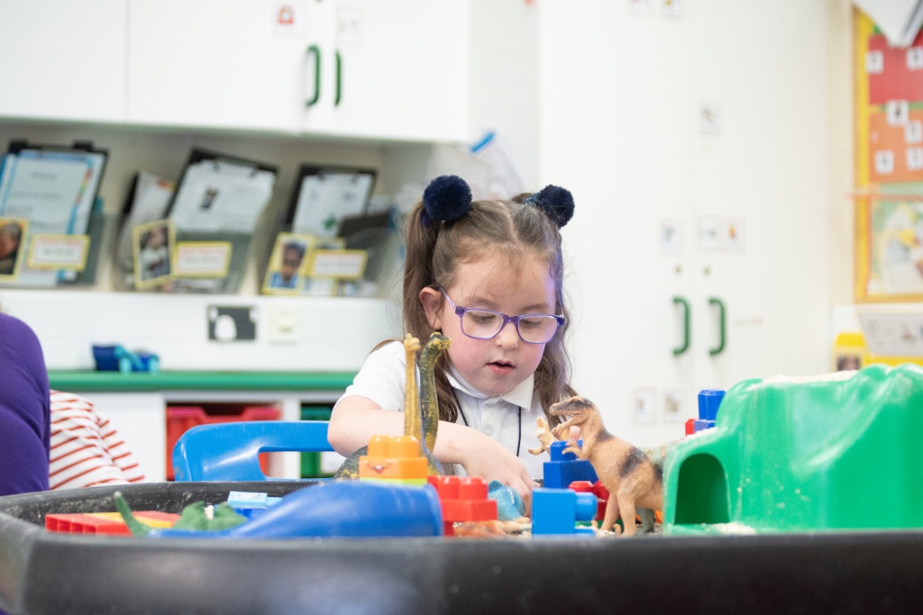 A young girl is seen sat a table, playing with Dinosaur toys inside a sandpit.