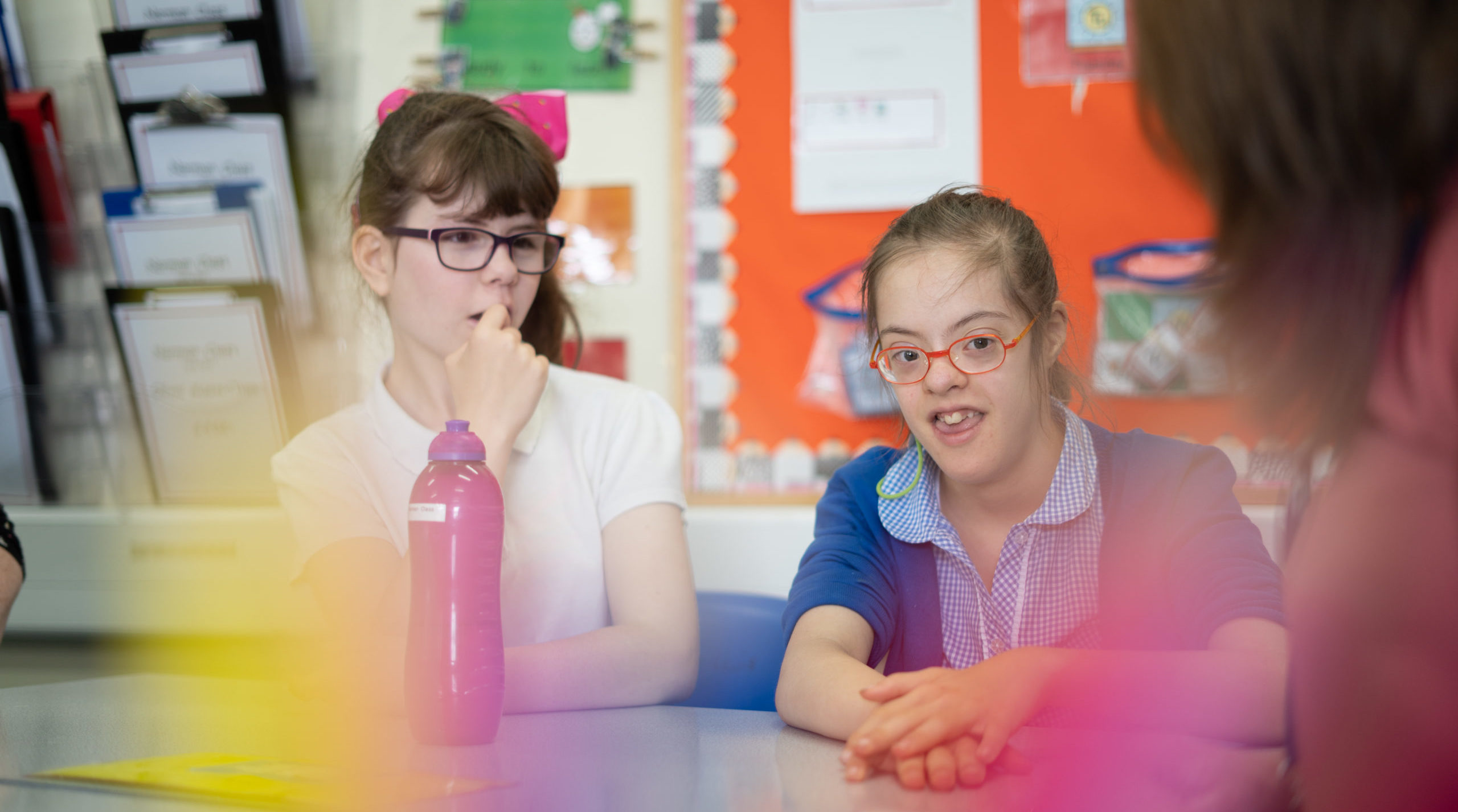 Two girls pictured sat at their desk, listening to the teacher speak to them inside a classroom.