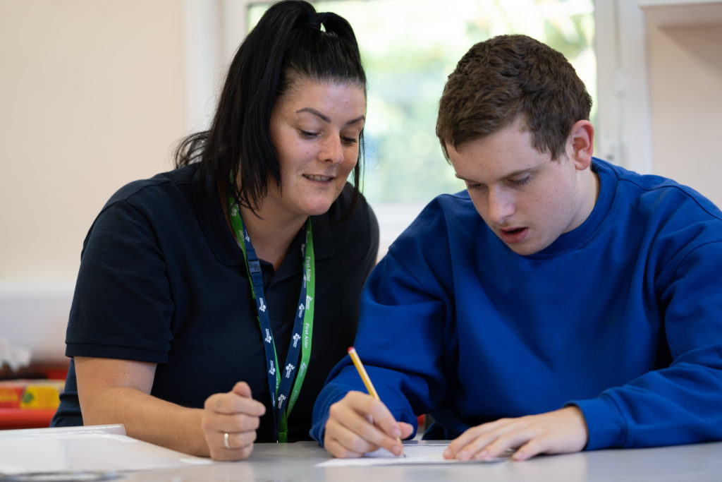 An older student is seen writing with a pencil on a piece of paper, assisted by a female staff member to his right.