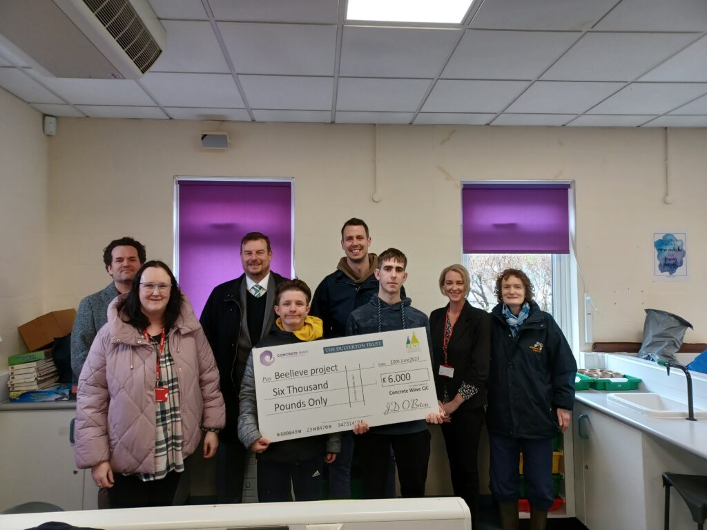 Students are seen welcoming visitors to the academy from the Beelieve Project. Two students can be seen holding a large cheque with a charity donation written on.