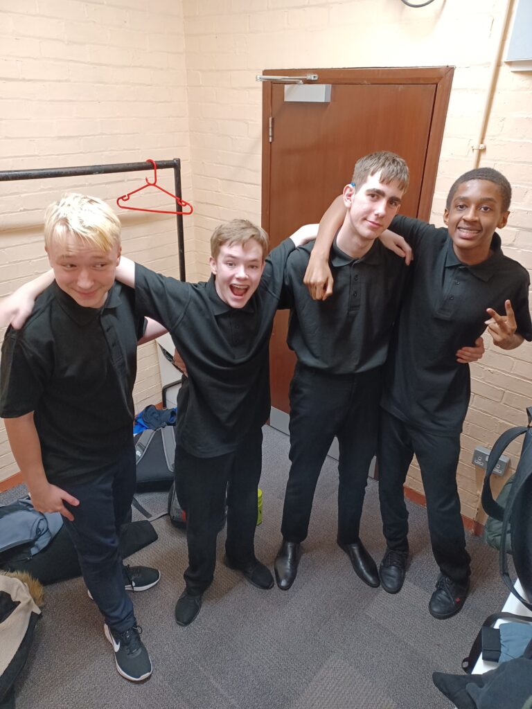 Four male students are seen posing for the camera together and smiling, backstage at the 2023 Coram Shakespeare Schools Festival.