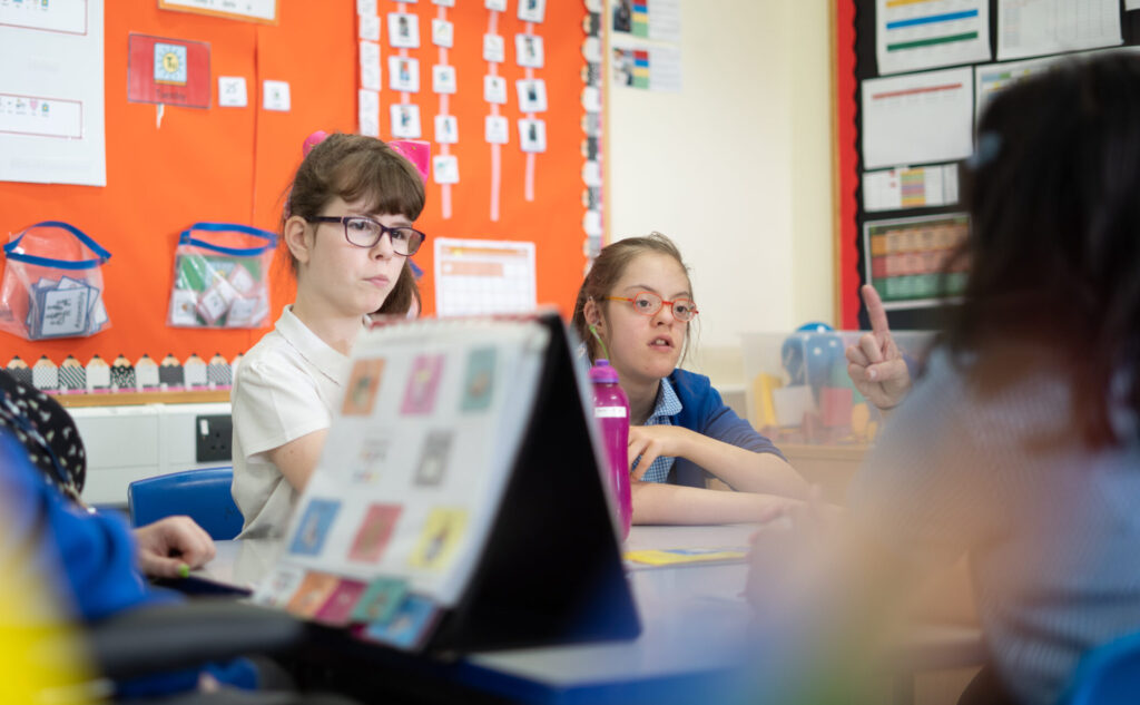 Two young girls are pictured sat next to each other at their desk, paying close attention to the member of staff sitting opposite them.