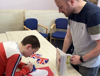 A male member of staff is seen assisting a pupil to create a replica of a postage stamp featuring the image of King Charles III on coloured paper.