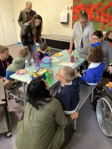 A group of students are shown having lunch together at a dinner table, under the supervision of staff and parents.