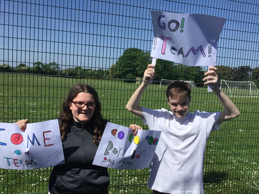 Two students, a boy and a girl, are pictured smiling for the camera, whilst holding up signs cheering on the student Football team.