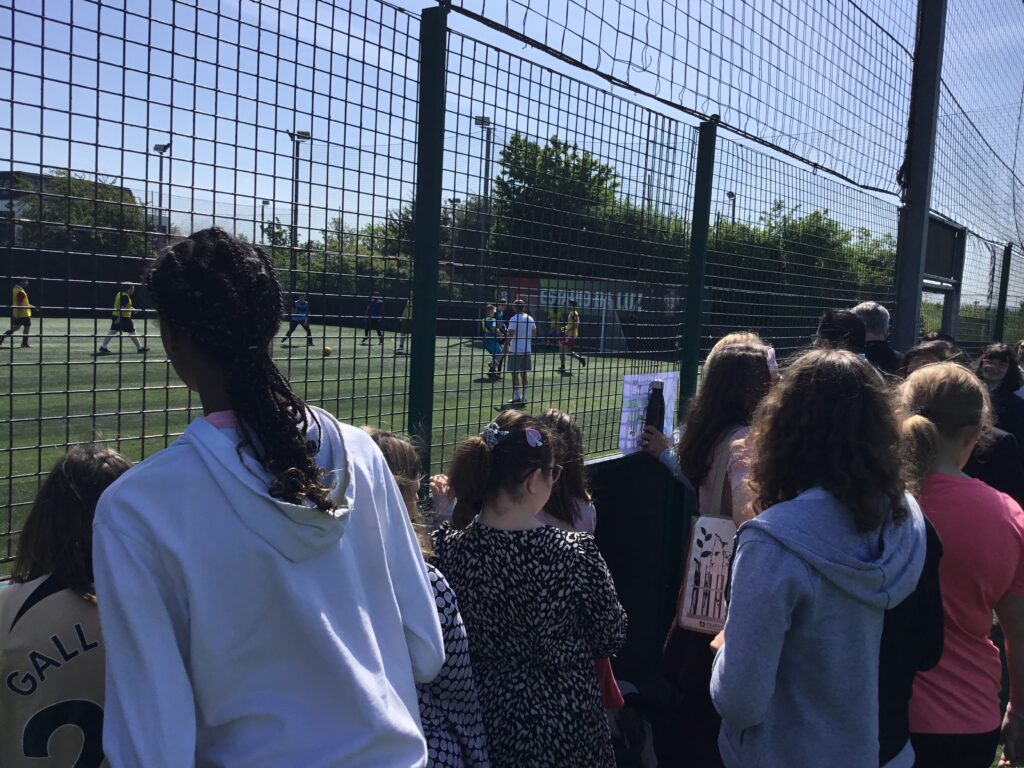 A large group of students are seen gathered around the astro turf fence to watch the team play a match of Football.