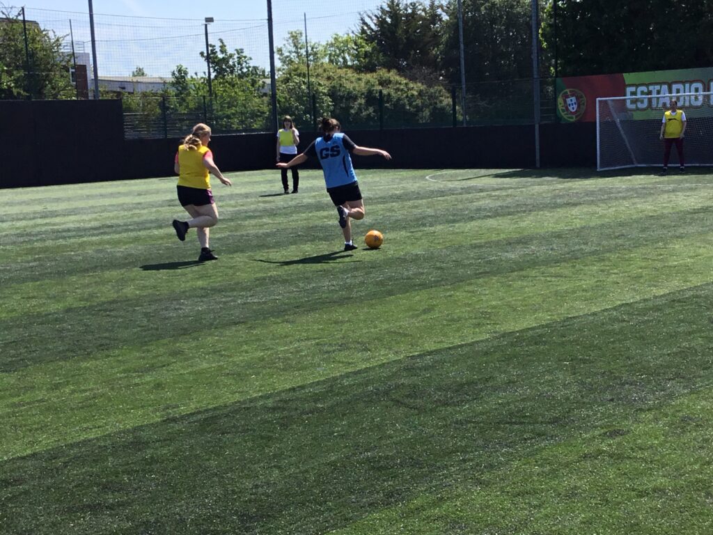 A team of students are pictured playing Football together, outdoors on an astro turf on the academy grounds. A girl in a blue vest is about to strike.