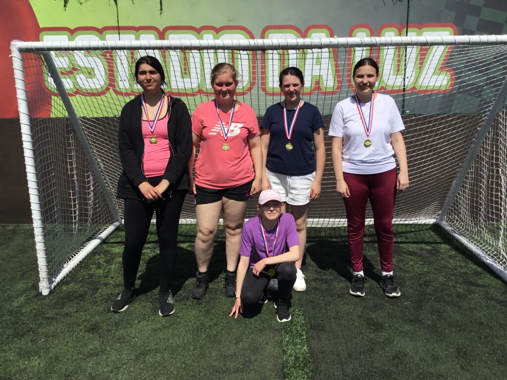 Five female students are pictured smiling for the camera underneath a goalpost, whilst wearing medals they have won for Football around their necks.