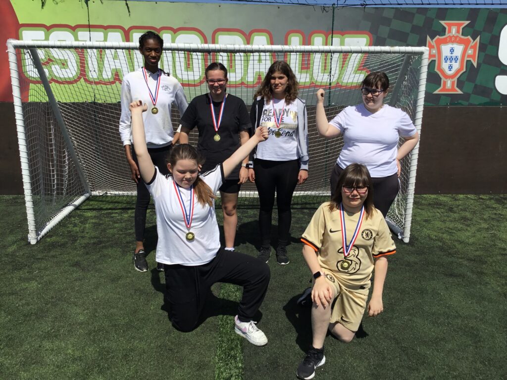 Six students are pictured smiling together for the camera, under a goalpost having just won medals for their Football success.