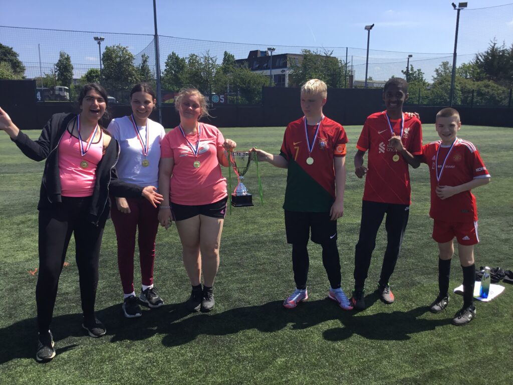 A group of students are seen celebrating together outdoors on the academy grounds, having just won a trophy for their Football success.