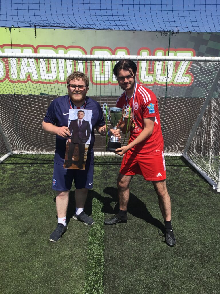 Two students are pictured celebrating together and smiling for the camera under a goalpost, after having won a trophy for their Football success.