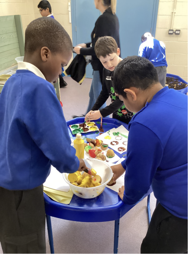 Three young pupils are pictured stood around a circular blue table, using craft materials and paint to create some artwork.