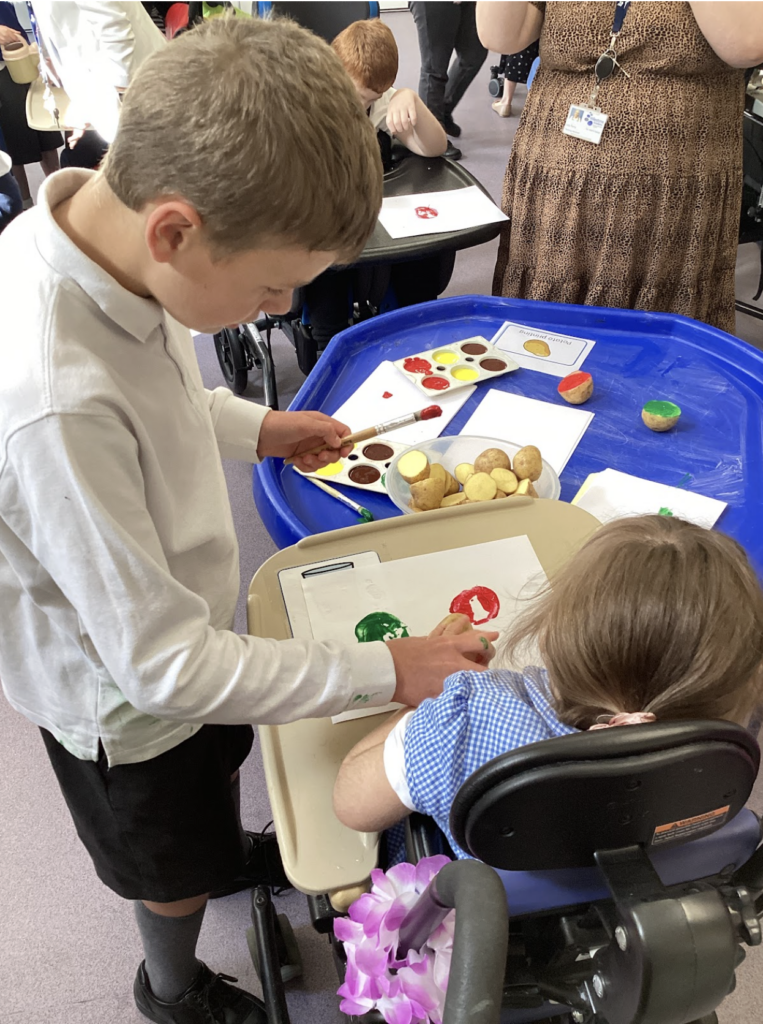 An older male pupil is pictured helping a younger female pupil in a wheelchair to create some artwork using paints.