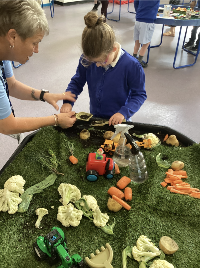 A young female pupil is pictured playing with a toy tractor at a desk with various vegetables spread across the table, imitating a farm scene. A female member of staff is seen supervising her as she plays.