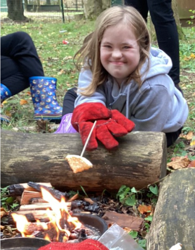 A young girl is seen smiling for the camera, whilst leaning forward to toast a marshmallow on a stick over a fire in the forest.