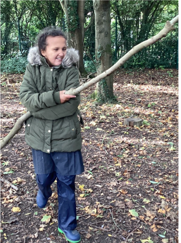 A female student is seen smiling for the camera, whilst outdoors in her winter coat and holding onto a long tree branch.