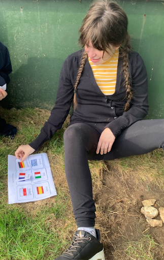 A female student is pictured sat on the grass with her back against a wall, looking at a sheet of paper with the flags from different countries around the world on it.