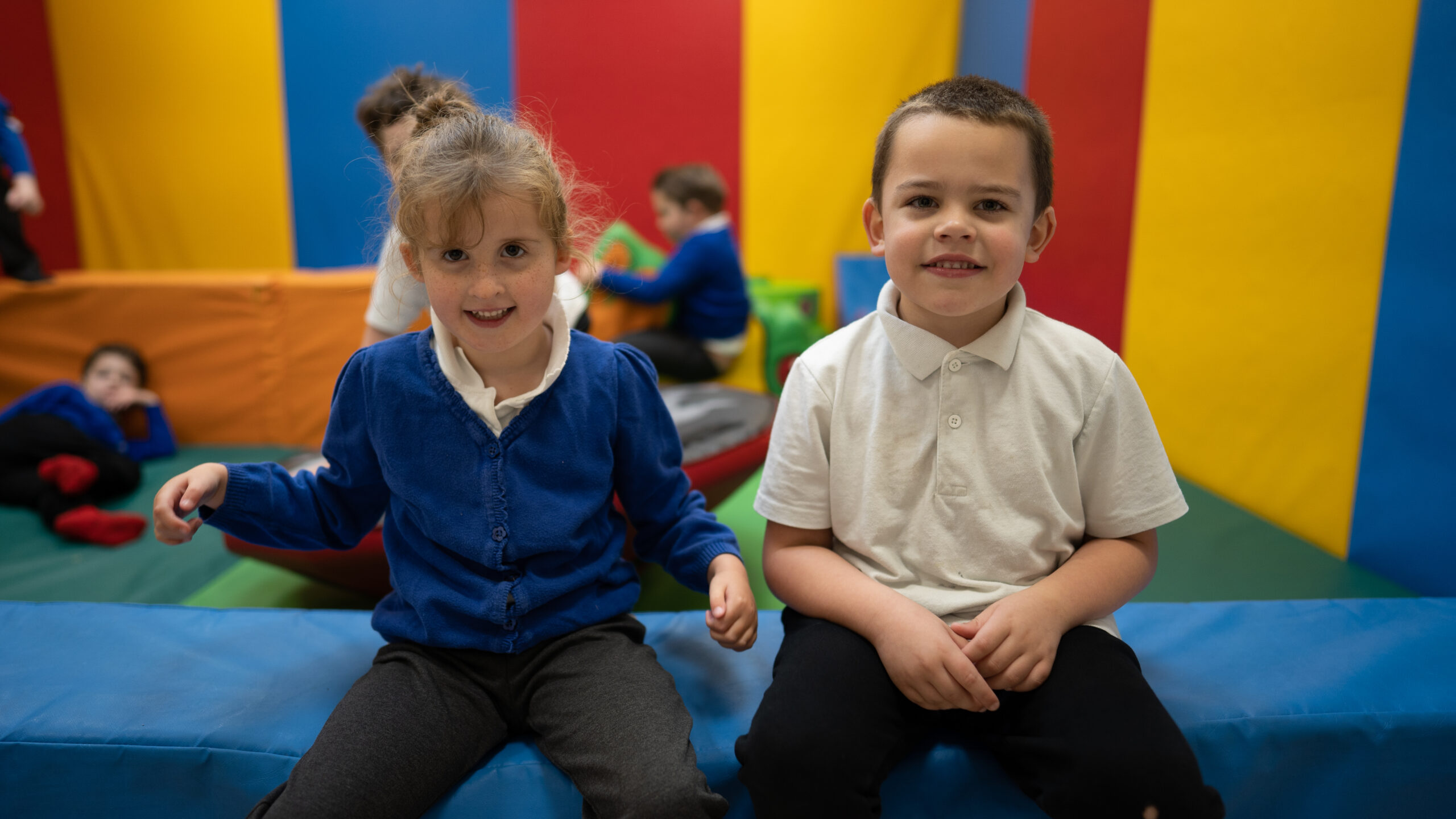 Two young children sitting in a soft play area smiling for the photo